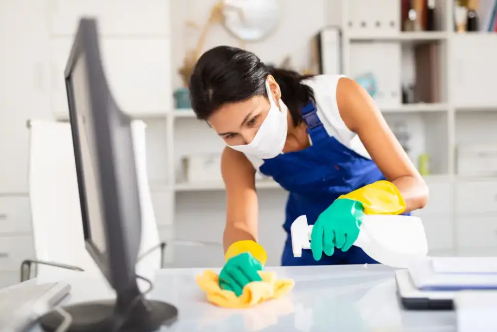 A maid is cleaning a computer desk with a cloth and a spreyer.